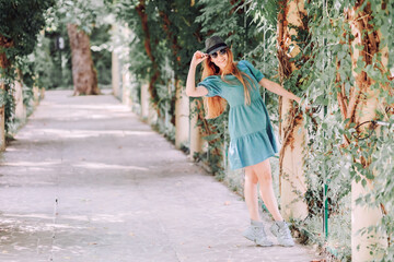 A girl in a blue dress posing while standing on tiptoes holding her hand to a metal bar of a lattice on an alley in a green park