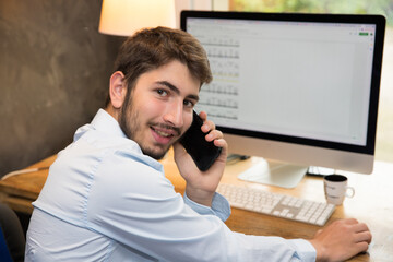 Jeune homme en télétravail ou bien étudiant devant un ordinateur en train de téléphoner avec son smartphone