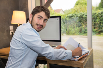 jeune homme au travail ou en télétravail devant un ordinateur en train d'écrire et regardant...