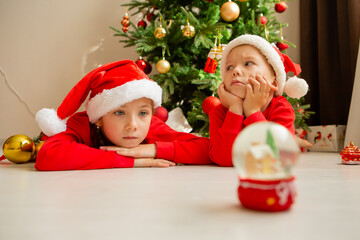 Obraz na płótnie Canvas two children, brother and sister in red Christmas pajamas look at a glass ball with a Christmas tree and a house inside, dream and make wishes. new Year's winter concept. celebrating at home