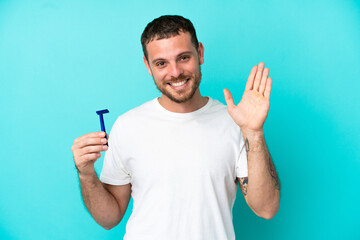 Fototapeta premium Brazilian man shaving his beard isolated on blue background saluting with hand with happy expression
