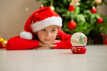 cute little girl in a red Christmas costume looks at a glass ball with a Christmas tree and a house inside and makes a wish. new Year's winter concept. celebrating at home. space for the text