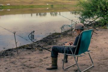 A boy with a spinning rod is fishing. Bank of a river or lake on a summer morning. Smartphone for social networking.