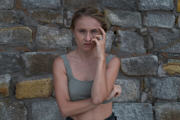 Sad pensive face of young caucasian blonde woman in crop top on the stone background. Young woman crying and holding her hand near the face, sad