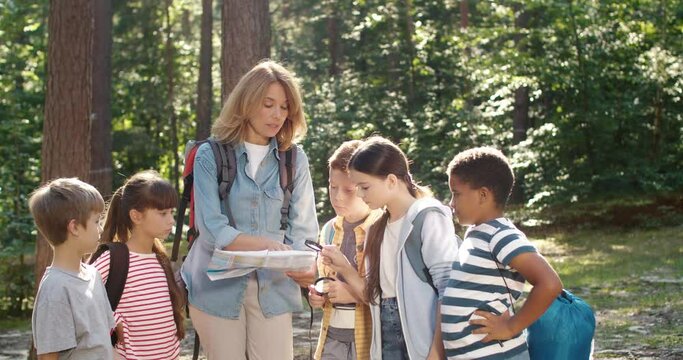 Caucasian woman teacher walking in forest and examining route plan with her pupils. Woman resting in wood with kids while having walk outdoor and looking at map. Tourism concept