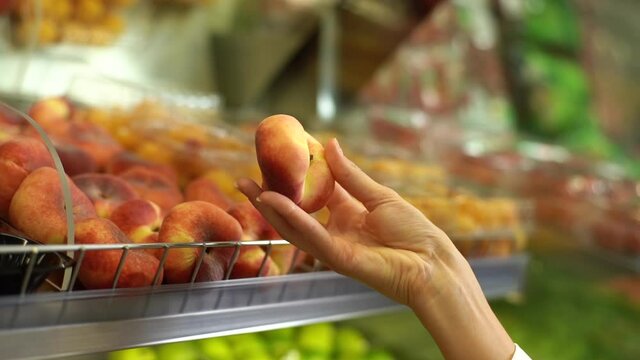 Close-up unrecognizable female hands standing by fruit stand at market and looking at fig peach. Closeup of young woman buyer picking peaches in supermarket. Lady decision buying nutrition fresh food.