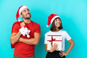 Young couple with christmas hat handing out gifts isolated on blue background looking up while smiling