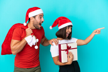 Young couple with christmas hat handing out gifts isolated on blue background pointing to the side to present a product