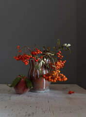 In a ceramic vase, there are rowan branches with bunches of red berries. Nearby lies a ripe peach with leaves. Still life.