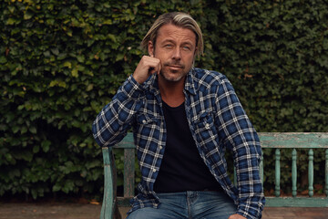 Fototapeta premium Man with blond hair and a stubble beard in a blue checkered shirt sits on a wooden bench in the backyard in summer.