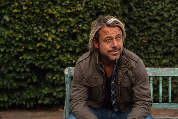 Fototapeta premium Smiling man with blond hair and a stubble beard in a green coat sits on a wooden bench in the backyard in summer.