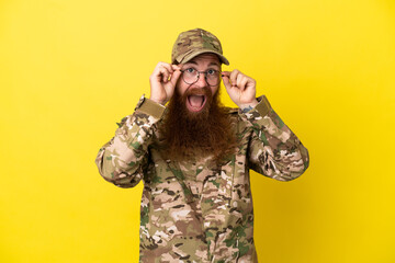 Military Redhead man over isolated on yellow background with glasses and surprised