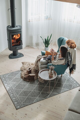 Young woman in a cozy armchair and a plaid sitting by the fireplace with a domestic cat, drinking...