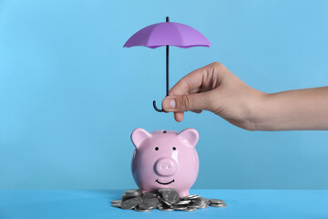 Woman holding small umbrella over piggy bank with coins on light blue background, closeup