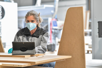 Woman reading technical informations on her tablet in a  industrial carpenter workshop and wearing protections against coronavirus
