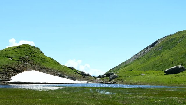Piles of snow, firn, at the foot of a green valley. Two pasture slopes in summer. View of a mountain lake.