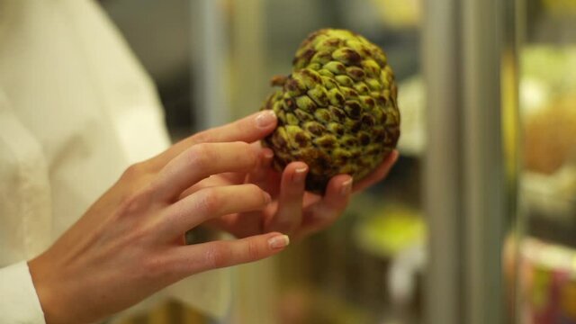 Close-up hands of unrecognizable young adult woman holding unknown exotic fruit and looking it. Female client holding small fresh ashta in hands at grocery store. Shooting in slow motion.
