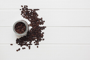 Top view of coffee beans in a jar, with some other beans scattered around it in a white wooden background