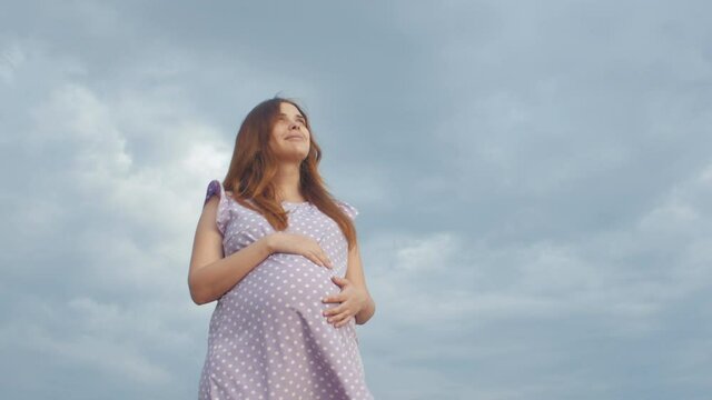 dreaming pregnant woman looks up to sky with gratitude in field on background of beautiful cloudy sky