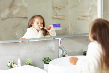 Obraz na płótnie Canvas A happy little girl in a robe brushes her teeth in front of a mirror in the bathroom. Morning routine. Hygiene.