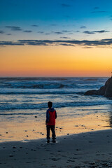 Naklejka premium A young Asian boy in red jacket walking along the shore of Rialto Beach during sunset. 