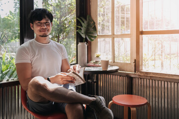 Happy Asian man drink coffee and reading book in warm light of summer.
