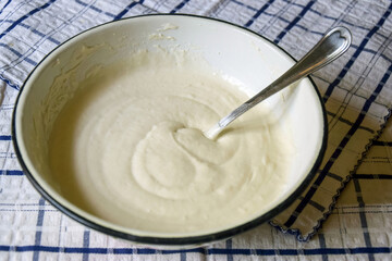On kitchen table there is an enameled bowl with spoon and liquid dough for baking sweet pie. Close-up. Photography, concept.