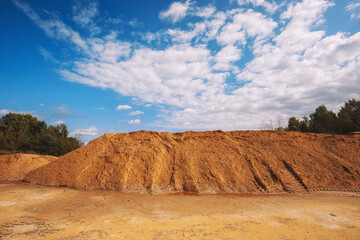 Large pile of industrial sand, stockpiled and prepared for use in construction and road construction, against a backdrop of blue sky and green forest