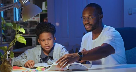 African american businessman father teaching and helping son doing homework in living room at late night. home studying at distance learning, Family spending quality time together. Lifestyle concept. - Powered by Adobe