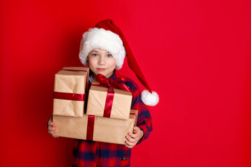 Happy holiday and Merry Christmas. Portrait of a boy in a cap with gifts in his hands on a red background. A place for text.