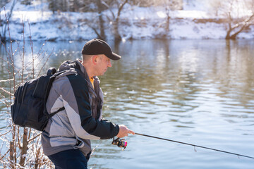 A fisherman with a fishing rod and a backpack catches fish on the bank of a snow-covered river in early spring