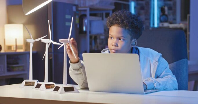 Young smart african american boy using computer learn about how wind turbines work from home at late night. Kids learning about eco-friendly forms of renewable energy. Technology and education.