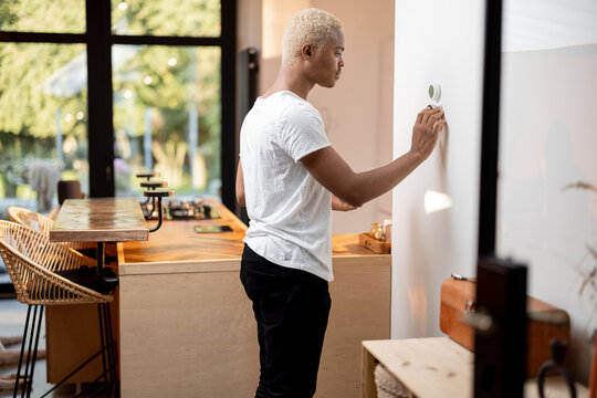 Black Man Choosing Temperature On Thermostat. Young Focused Guy Pushing Button On Smart Home System. Concept Of Modern Domestic Lifestyle. Interior Of Kitchen In Modern Apartment.