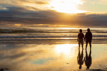 Fototapeta premium A mother and son travelling together along the Cannon Beach in Oregon looking outwards towards the horizon during a golden sunset