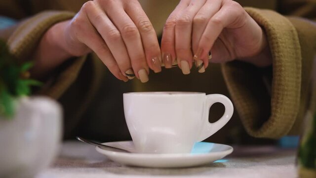 Girl opens stiker of sugar and pours granules of sweetener into cup with drink. Woman mixes tea or coffee by teaspoon. Harmful carbohydrates - glucose. Fragile fingers of girl and ceramic cup close up
