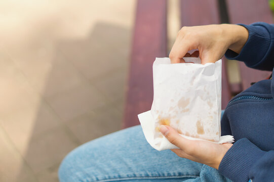 A Young Girl's Hand Takes Out Slices Of French Fries From A White Paper Bag. A Hipster Girl Eats Her Delicious French Fries Right On The Street, Sitting On A Bench In A City Park. Fast Food. Junk Food