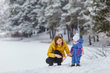 Fototapeta na wymiar Young woman sculpt snowballs with her baby girl in the winter forest. Cute kid in warm clothes having fun outdoor with mother. Mom and daughter. Family holidays leisure, motherhood, love in family