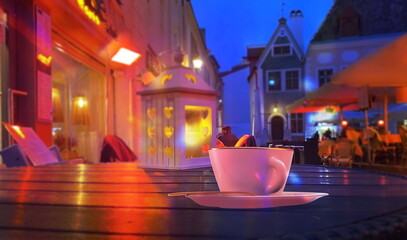 neon night city blurred light street restaurant yellow coffee cup lantern on wooden table medieval house in Tallinn Old Town Estonia, vintage lantern at night street in festive medieval Tallinn Old To