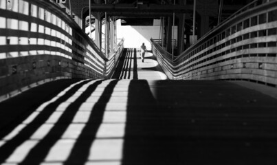 A black and shite image person walks across a city bridge in the shadows
