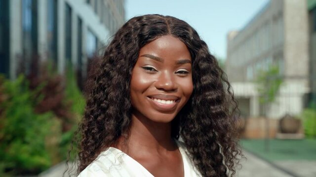 Happy smiling young ethnic black African attractive millennial business woman or student with curly long hair standing outdoor on sunny city street looking at camera. Slow motion close up portrait.