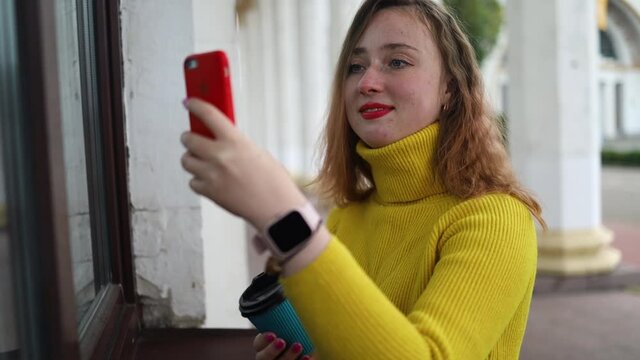 young blonde woman is wearing a yellow sweater, She is holding a coffee to go and taking selfie by smartphone.