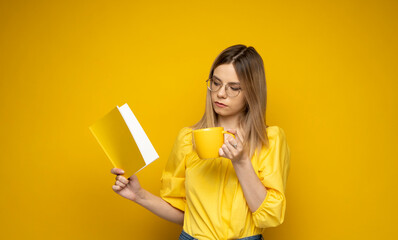 Beautiful smart young girl holding and reading book isolated on the yellow background. Portrait of attractive woman in a yellow blouse and wearing glasses reading book. Education, studying, knowledge.