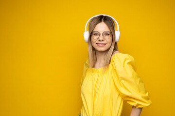 Beautiful attractive young blond woman wearing yellow t-shirt and glasses in white headphones listening music, dancing and laughing on blue background in studio. Relaxing and enjoying. Lifestyle.