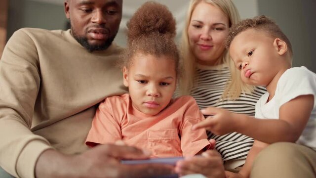 Interested multiracial family of four using smartphone together at home. Biracial kids and diverse parents playing mobile video game, browsing Internet or looking through pictures on cell phone