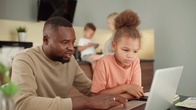 Panning shot of Black man working on laptop from home or browsing Internet and talking with cute biracial little daughter. Father and daughter using pc. Wife and toddler son in background