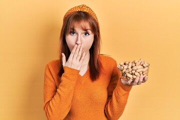 Redhead young woman holding peanuts covering mouth with hand, shocked and afraid for mistake....