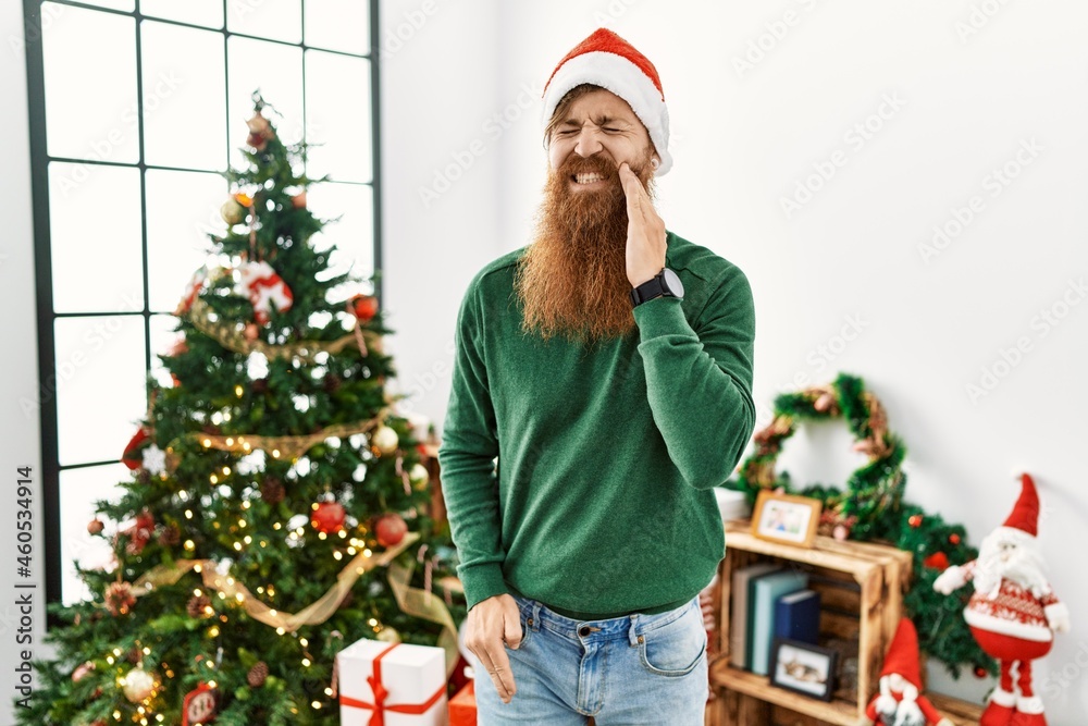 Wall mural Redhead man with long beard wearing christmas hat by christmas tree touching mouth with hand with painful expression because of toothache or dental illness on teeth. dentist