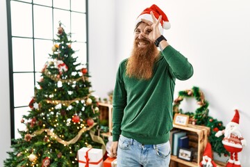 Redhead man with long beard wearing christmas hat by christmas tree doing ok gesture with hand smiling, eye looking through fingers with happy face.