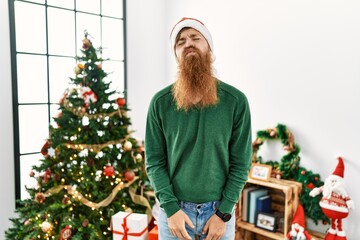 Redhead man with long beard wearing christmas hat by christmas tree looking at the camera blowing a kiss on air being lovely and sexy. love expression.