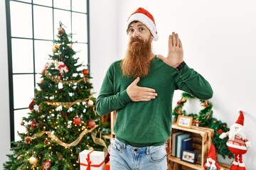 Redhead man with long beard wearing christmas hat by christmas tree swearing with hand on chest and open palm, making a loyalty promise oath